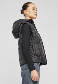 Schwarze, gesteppten Weste mit Kapuze, mit einem Reißverschluss vorne und seitlichen Taschen, getragen über einem langen schwarzen Oberteil. Weiche, gepolsterte Textur.
