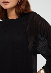 Zwarte top met geplooide textuur, voorzien van doorschijnende mouwen en een ronde halslijn. De gladde stof contrasteert met de geribbelde details.