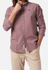 Camisa a cuadros roja y blanca de botones, con mangas largas, que presenta un logo azul en el pecho, combinada con pantalones beige claros.