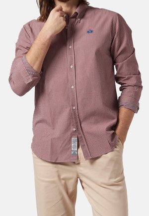 Camicia a quadri rossi e bianchi con maniche lunghe, caratterizzata da un logo blu sul petto, abbinata a pantaloni leggeri beige.