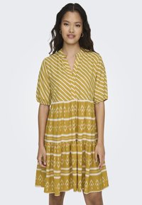 Senfgelbes Kleid mit kurzen Puffärmeln, vertikalen und horizontalen weißen Streifen, gestuftem Rock und tribal-inspirierten Mustern.