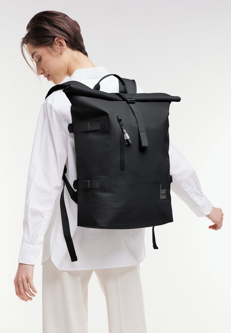 Persona con una gran mochila negra de tipo roll-top con bolsillo frontal con cremallera y correas laterales, vistiendo camisa y pantalones blancos, de perfil.