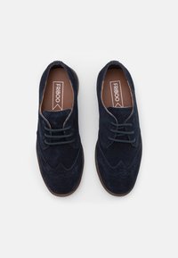 Scarpe in suede blu navy con un design classico a lacci, caratterizzate da dettagli wingtip e una suola in gomma marrone. Upper testurizzato con cuciture a contrasto.