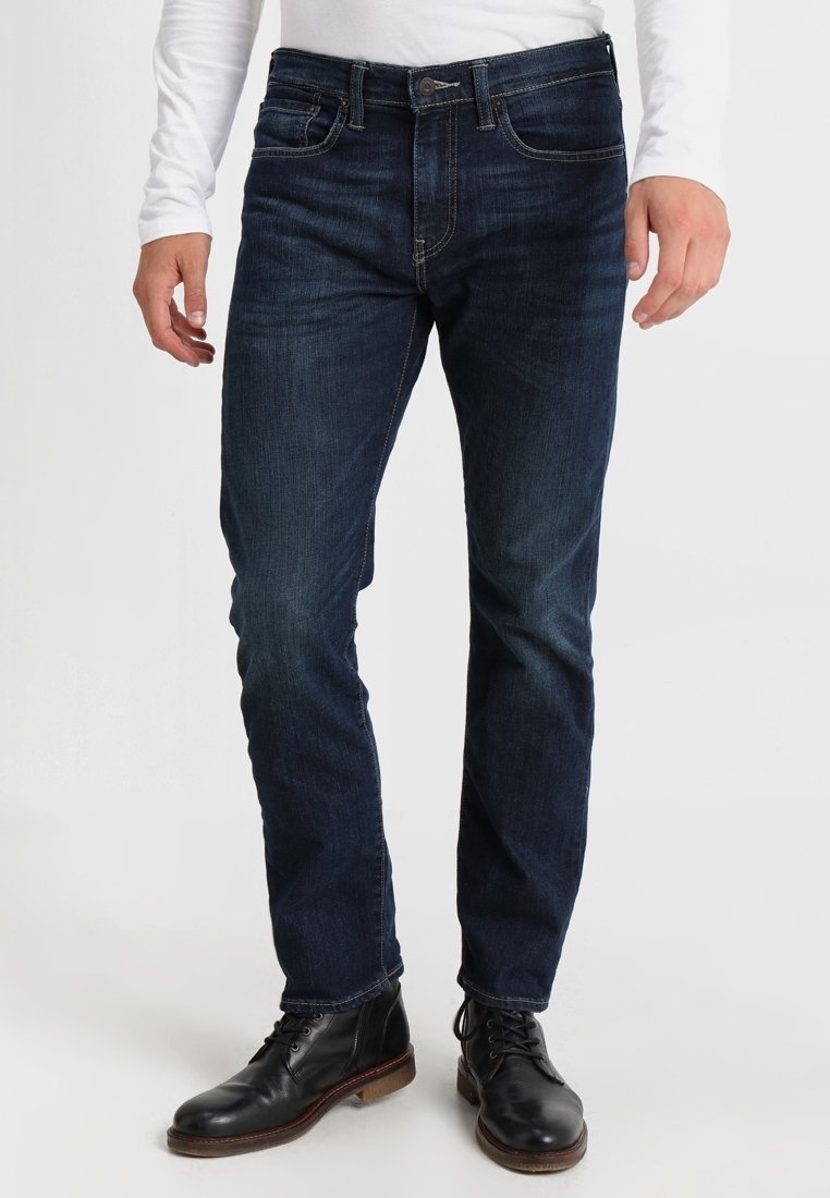 Homme portant un jean bleu foncé coupe droite, une chemise blanche à manches longues et des bottes noires à lacets, debout devant un fond uni.