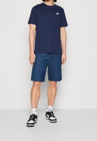 Marinblå bomullströja med rund halsringning och vit Nike-logga; tillsammans med denimshorts som når knäna och svart-vita sneakers.