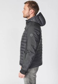 Zwarte hybride jas met een gequiltte voorkant, gladde achterkant, verstelbare capuchon en ribgebreide manchetten; bevat een ronde logo patch op de mouw.