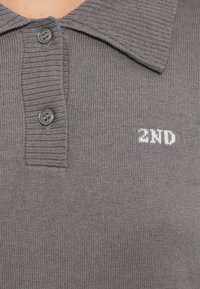 Szürke kötött póló, bordázott gallérral, két gombbal és texturált felülettel, amelyen fehérrel hímzett "2ND" felirat található.
