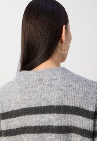 Femme aux longs cheveux foncés portant de grandes boucles d'oreilles argentées en forme d'anneau et un pull gris en tissu duveteux avec deux bandes horizontales noires dans le dos.