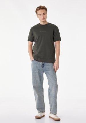 Jonge man staand met handen in de zakken, draagt een donkergroen T-shirt met de tekst "OWN YOUR PATH", lichtblauwe spijkerbroek en witte sneakers.