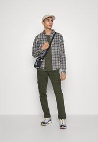 Camisa a cuadros en verde, beige y azul marino, combinada con una camiseta verde oliva y pantalones cargo. Una gorra beige y una pequeña bolsa negra acentúan el conjunto.