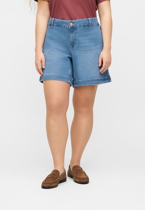 Personne portant un short en jean retroussé, des chaussures marron sans lacets et un haut bordeaux à manches courtes, debout devant un fond clair uni.