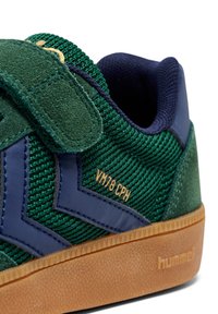 Sneaker verde con texture, con accento in pelle blu, cinturino in velcro e suola in gomma color naturale. Dettaglio logo dorato vicino al tallone.