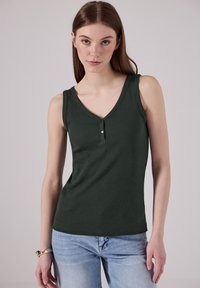Donna con la pelle chiara e lentiggini indossa un top a maniche corte di colore verde scuro a coste con dettagli di bottoni e jeans azzurri su uno sfondo semplice.