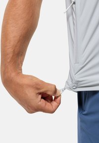 Maglietta sportiva grigia chiara con lacci all'orlo, dotata di una tasca laterale con zip. Mano che stringe il laccio per la regolazione.