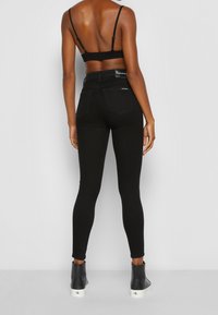 Mujer vestida con jeans skinny de tiro alto negros, un bralette negro y zapatillas altas negras, de pie contra un fondo gris liso.