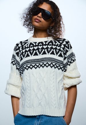 Άτομο που φοράει υπερμέγεθες μαύρα γυαλιά ηλίου και λευκό πουλόβερ πλεκτό με μαύρα γεωμετρικά σχέδια, με τα χέρια στις τσέπες ενός μπλε τζιν.