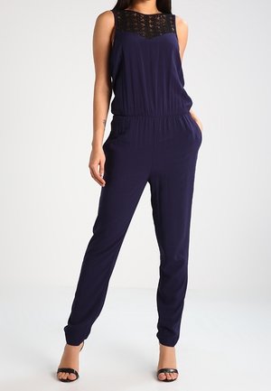 Femme portant une combinaison sans manches bleu marine avec un détail en dentelle au niveau du décolleté, une taille élastique, des jambes fuselées et des talons noirs ouverts à l'avant.