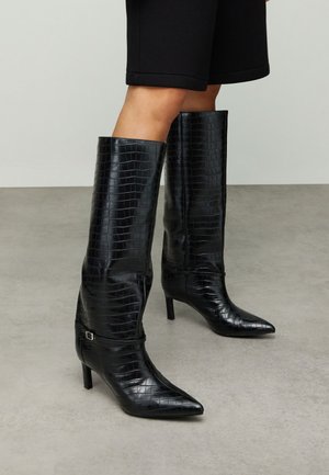 Schwarze kniehohe Stiefel aus krokogeprägtem Leder mit spitzer Schuhspitze, schlanker Silhouette und kleinem Schnallenakzent am Knöchelriemen.