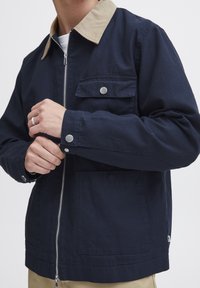 Mörkblå jacka med beige krage, dragkedja fram och fickor. Har silverfärgad detaljering och en avslappnad passform. Tillverkad av bomull med en mjuk textur.