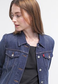 Mørkeblå denimjakke med klassisk krave, frontlommer, metalknapper og brune sømdetaljer. Brukt over en svart topp.