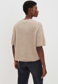 Persona che indossa un maglione fuzzy beige a maniche corte e jeans neri, volgendo di lato su uno sfondo bianco semplice.