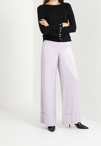 Zwarte blouse met lange mouwen en ruche-mouwen, versierd met witte bloemen borduursels, gecombineerd met lichtpaarse broek met wijd uitlopende pijpen en accent op de zoom.