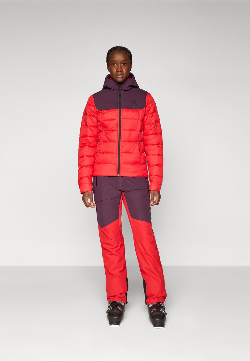 Czerwona i ciemnofioletowa ocieplana kurtka z zamkiem błyskawicznym i kapturem, zestawiona z dopasowanymi spodniami narciarskimi z kątowym blokowaniem kolorów i zamkami.
