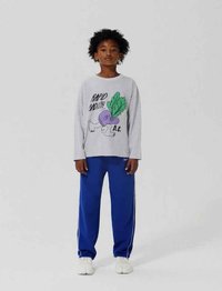 Child with curly hair stands wearing a gray long-sleeve shirt with a radish graphic and blue pants against a plain background.