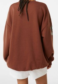 Sweat-shirt marron surdimensionné avec un col rond et des épaules tombantes. Il présente un motif étoile sur la manche gauche et des poignets côtelés.