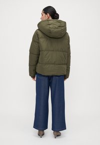 Vrouw met donker haar in een knot, gekleed in een olijfgroen pufferjack met capuchon, wijd uitgesneden blauwe jeans en taupe hoge hakken, staande met de rug naar de kijker.