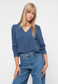 Blå V-halsbluse med lange, rynkede ærmer, parret med højtaljede, lyse denimjeans. Glat tekstur og afslappet pasform.