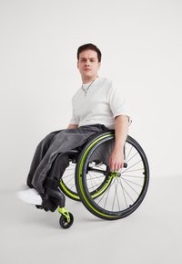 Czarny wózek inwalidzki z neonowozielonymi akcentami, wyposażony w grube koła i wyściełane siedzisko. Osoba ma na sobie białą koszulę i szare spodnie.