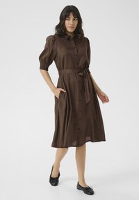 Robe marron à boutons avec des manches courtes à volants, une ceinture à la taille et des poches latérales, fabriquée en tissu léger et lisse. Chaussures plates noires.