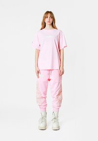 Camiseta de algodón rosa con un parche rectangular, combinada con pantalones de chándal rosa que tienen acentos bordados en los laterales y puños elásticos, llevados con botas blancas.