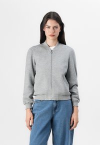 Ung kvinne som står mot en hvit bakgrunn, iført en grå bomberjakke med glidelås og løse blå jeans, med rett hår og en nøytral uttrykk.