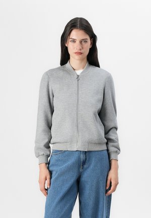 Jeune femme se tenant devant un fond blanc, portant une veste bomber zippée grise et un jean bleu ample, avec des cheveux raides et une expression neutre.