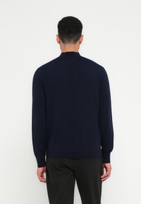 Temno modra pletena svetilka z dolgimi rokavi, visokim ovratnikom in rebrastimi manšetami. Gladka tekstura, oprijet silhueta in minimalističen dizajn.