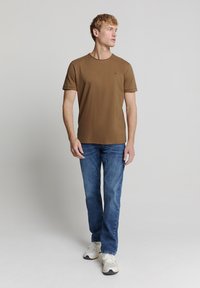 Bruine short-sleeve t-shirt met ronde hals, gecombineerd met lichtblauwe spijkerbroek en witte sneakers, gepresenteerd op een staande mannelijke model.