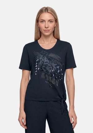 Femme aux cheveux blonds et raides, portant un t-shirt noir à manches courtes avec un motif floral et du texte, associé à un pantalon noir sur fond uni.