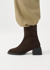 Brown suede ankle boot with a pointed toe and a chunky black heel, paired with beige, gathered trousers.