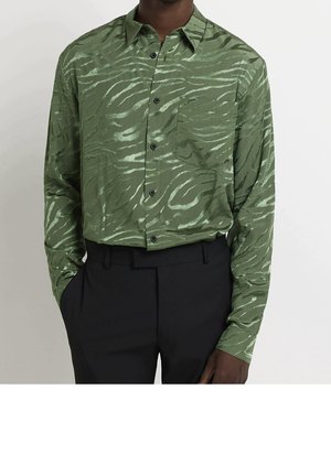 Mand iført grøn langærmet skjorte med mørkere tigerstribet mønster, stoppet ned i sorte bukser, hånd i lommen, på lys baggrund.