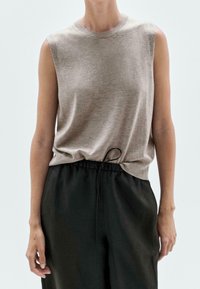 Beige ilmanhihaisa toppi, joka on valmistettu pehmeästä kankaasta, jossa on pyöreä pääntie ja lievää laskostusta helmassa, yhdistettynä väljiin mustiin housuihin.