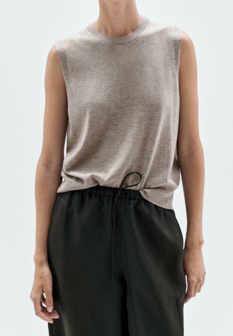 Beige ilmanhihaisa toppi, joka on valmistettu pehmeästä kankaasta, jossa on pyöreä pääntie ja lievää laskostusta helmassa, yhdistettynä väljiin mustiin housuihin.