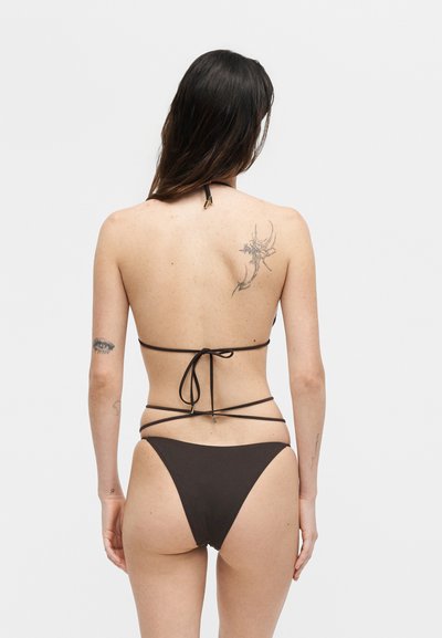 Γυναίκα με μακριά σκούρα μαλλιά που φοράει μαύρο στρινγκ μπικίνι, δείχνοντας την πλάτη με λουλουδένια και ματιών τατουάζ στα μπράτσα και τον ώμο.