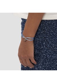 Bracelet en cordon bleu avec un accent argenté et un pendentif circulaire, porté au poignet, sur une jupe texturée foncée.