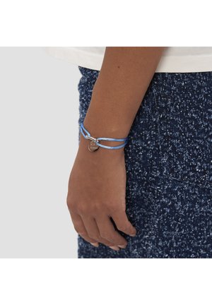 Blått snörearmband med en silveraccent och en rund berlock, bärs på en handled mot en texturerad mörk kjol.