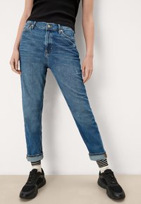 Modré denimové džíny s volným střihem, ohrnutými manžetami, pěti kapsami a minimálním opotřebením. Nosené s černými teniskami a pruhovanými ponožkami.