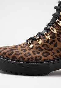 Stivale alla caviglia in pelle scamosciata leopardata marrone con dettagli in metallo dorato, lacci neri e una spessa suola in gomma nera. Presenta una punta arrotondata e cuciture.