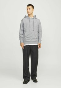 Grauer Kapuzenpullover mit Zugbändern, gerippten Bündchen und Saum, über einem weißen Hemd getragen, kombiniert mit weiten schwarzen Hosen und schwarzen Schuhen.