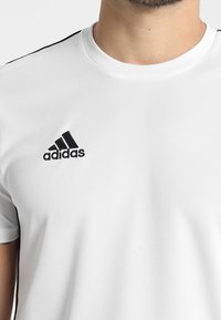 Bílý sportovní tričko s kulatým výstřihem, černé logo Adidas na levé straně hrudi a černé pruhy na ramenou. Mřížková textura.